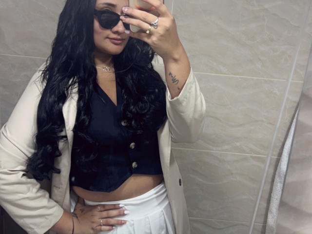 ElyRoss has long black hair, wears dark sunglasses, a black vest, white skirt, and beige jacket, with a visible tattoo on her right wrist, taking a mirror selfie.