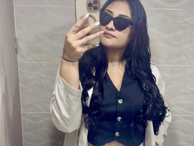 ElyRoss with long black hair, wearing black vest, white shirt, and dark sunglasses, taking a mirror selfie with a gold phone, showing white nails and a necklace.