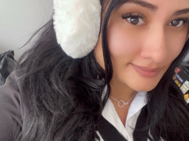 ElyRoss with long black hair, wearing fluffy white ear thingies, black vest, white shirt, and necklace, smiling at camera, brown eyes, subtle makeup, cute, soft expression.