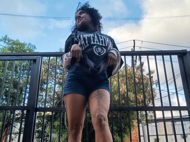 LunaKeller, com cabelos cacheados, vestindo uma camiseta preta com a inscrição "Satellite" e shorts azuis, em pé, exibindo uma tatuagem na coxa direita, olhando para baixo, em frente a uma cerca preta..