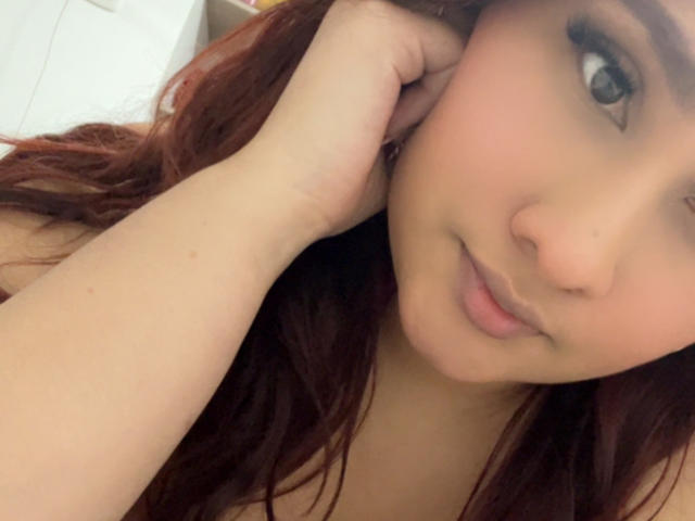 ElyRoss has red hair, brown eyes, and a cute face, with her hand near her ear, looking up with a slight smile. Her skin is light and her lips are pink.
