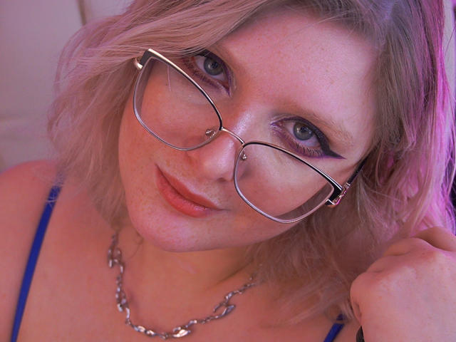Close-up of CoolTatis with light blonde, wavy hair and pinkish highlights. She has fair skin, blue eyes with purple eyeshadow, and a silver nose ring. She's wearing large, silver, rectangular glasses and a blue strap. Her lips are slightly parted with a subtle pink lipstick. She's holding her hair with her right hand, wearing a silver chain necklace. The lighting is pink and purple, giving a soft, warm glow to her skin. The image has a slightly warm, digital filter.