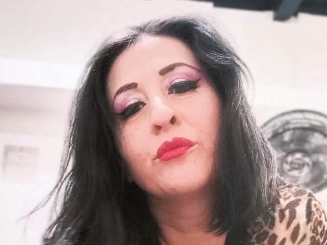 ScarletMilfX with black hair, pink eyeshadow, heavy eyeliner, red lipstick, and a leopard print top, pouting slightly. Her skin is pale, and she's looking down.