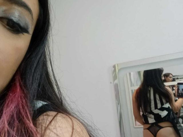 Close-up of RozarioPinceles, right eye and eyebrow visible with heavy black and silver eyeshadow. Partial face shows black hair with pink tips. Reflection in mirror shows RozarioPinceles from behind, wearing a black and white striped top, black thong, and long black hair. Mirror is slightly off-center, white wall behind. RosarioPinceles' back is mostly to the camera, showing thong and striped top. Slightly blurry, casual selfie style.