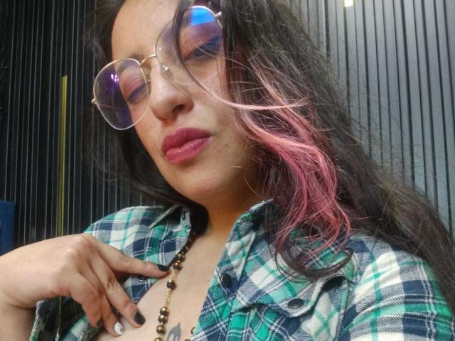 RozarioPinceles with pink-tipped hair, glasses, pink lipstick, green plaid shirt, necklace, and hand on chest, looking down. Boobs slightly visible under shirt. Soft, confident expression.