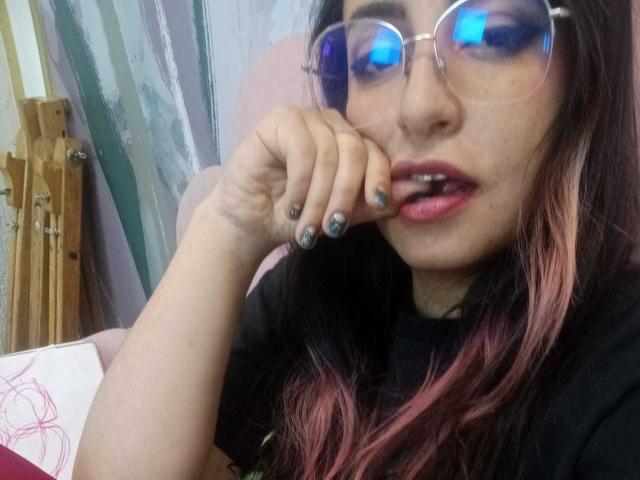 RozarioPinceles with blue reflective glasses, pink and black ombre hair, black shirt, pink lipstick, biting finger with grey nail polish, looking at camera.