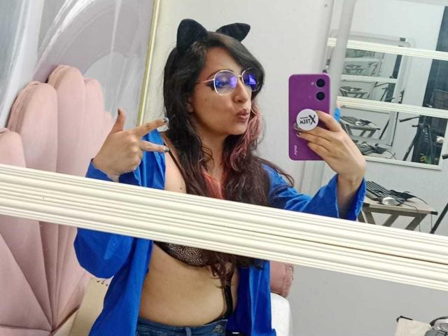 RozarioPinceles, wearing a blue open shirt, black bra, cat ears, and purple sunglasses, taking a mirror selfie with a purple phone, showing off her tits and pout.