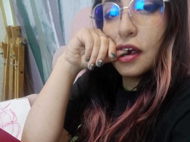 RozarioPinceles with pink-tipped hair, glasses reflecting blue light, black shirt, finger touching slightly open mouth, nails painted dark blue, looking seductive.