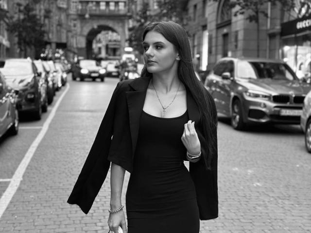 LisaDark with long dark hair, wearing a black dress and open jacket, standing on a cobblestone street, looking to the side, holding her jacket, with a necklace and bracelet.