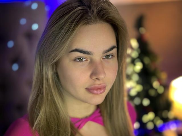LunaHeadly with blonde hair, pink top, and cute face, has full lips and a slight smirk. Her tits are small, and she's looking directly at the camera.