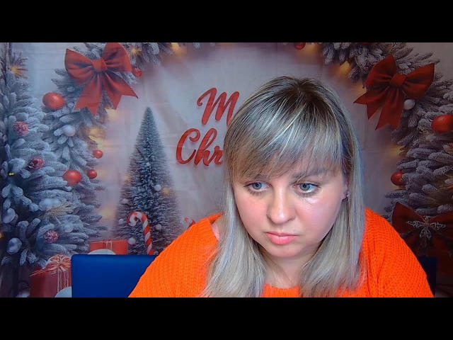 VeronicaSvitt has blonde hair, blue eyes, and a pouty face, wearing an orange sweater, with Christmas decorations in the background. Her tits are average, and she looks cute.