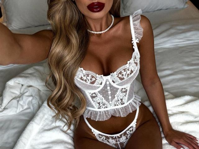 ElzaKomx with long, wavy blonde hair, wearing white lace lingerie with frilled edges, pearl necklace, and dark red lipstick, sitting on a white bed. Her tits are prominent.