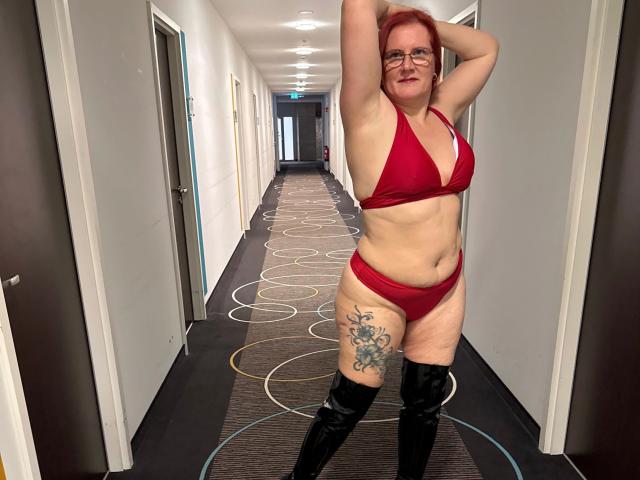 MrsEssen7471-hot with red hair, glasses, and a red bikini, showing a tattoo on her right thigh, wearing black thigh-high boots, arms raised, smiling in a hallway.