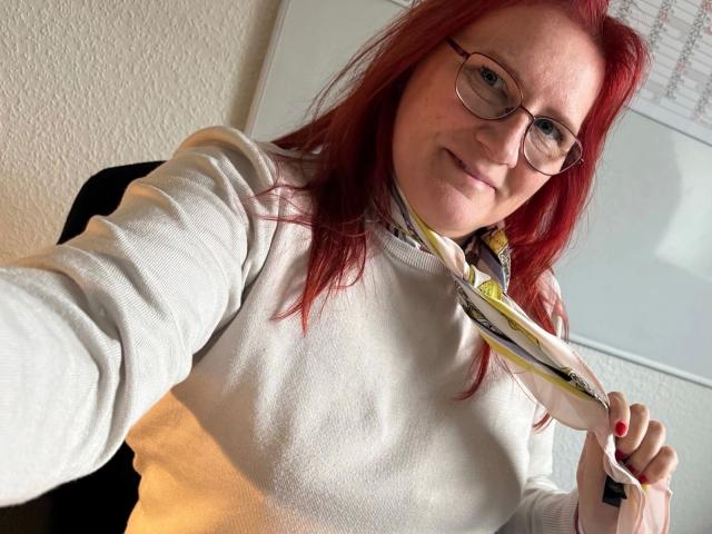 MrsEssen7471-hot with red hair, glasses, and a white sweater, holding a white phone charger, smiling, and taking a selfie, showing her small tits and cute face.