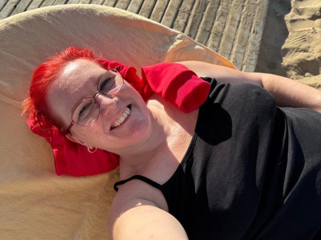 MrsEssen7471-hot with red hair, glasses, black tank top, and red pillow, smiling on a yellow beach chair, sunlit, showing her ass and tits slightly.
