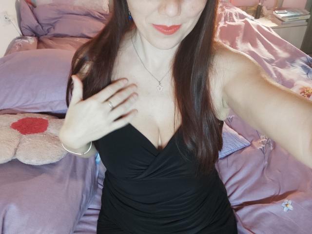 Cassy with long brown hair, red lipstick, black dress, showing her tits with a hand, wearing a necklace, on a bed with purple sheets.