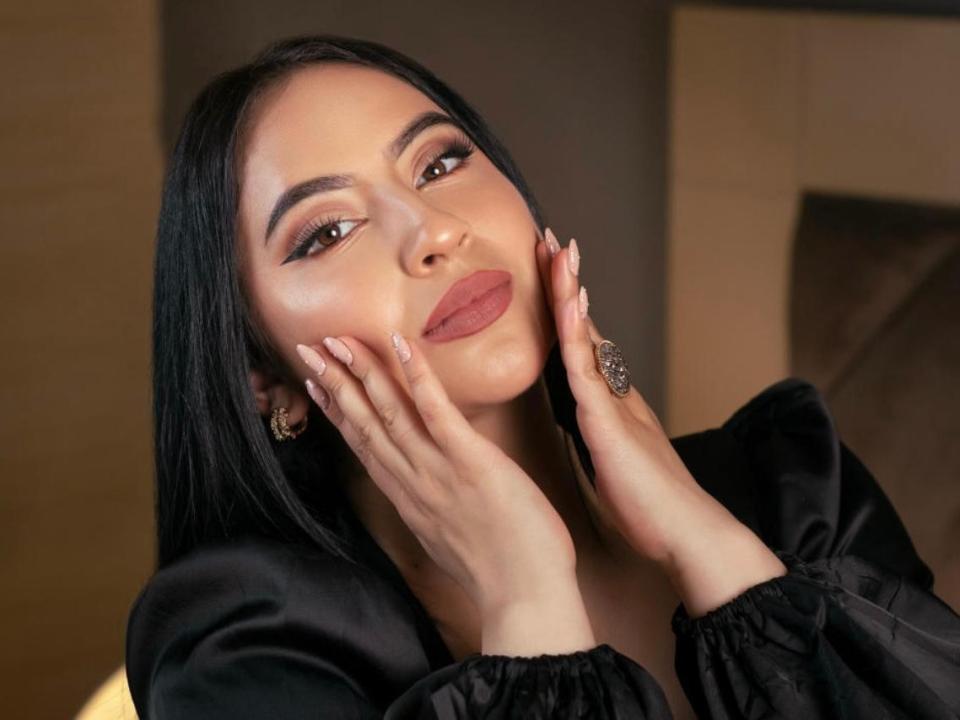 AbrilMontoya with black hair, hands on cheeks, wearing a black satin top, has full lips, brown eyes, and a ring on her right hand. Her ass is not visible.