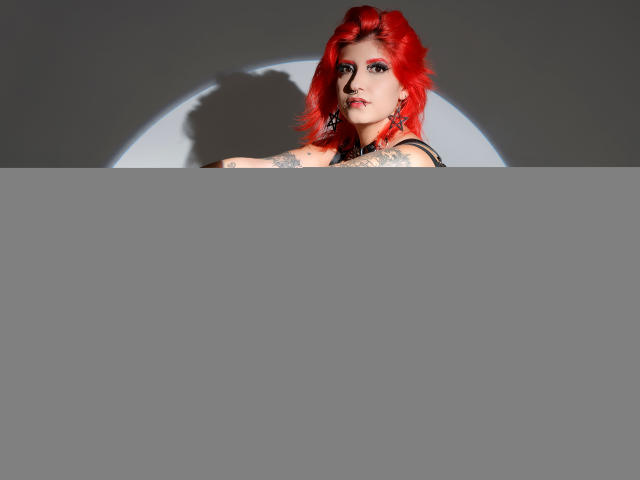 Athena Grant, con cabello rojo brillante, vestida con lencería negra de tirantes, sentada en un taburete, mostrando tatuajes en los brazos y el muslo, con un círculo sombreado detrás de ella..