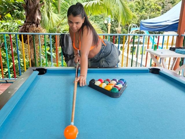 LissReyess, a woman with dark hair in a ponytail, leans over a blue pool table, gripping a cue stick with an orange tip. She wears a orange bra and blue jeans. A triangular pool ball rack with multicolored balls sits on the table's right. The background includes green foliage and a colorful metal railing. The composition emphasizes the pool table and LissReyess's focused posture. The image is clear and detailed.