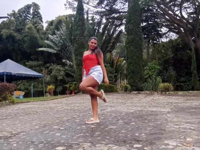LissReyess stands on a stone-paved area, mid-jump, with her right leg bent and left foot raised. She wears a red strapless top, light blue shorts, and white sandals. Her straight brown hair is shoulder-length, and she has a red lipstick. The background features lush green trees, a blue canopy tent, and various plants. The stone pavement is irregularly shaped, adding texture to the scene. The composition is centered, highlighting LissReyess in the foreground. The image has a casual, outdoor setting.