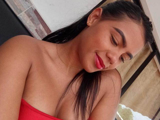 LissReyess has tan skin, long black hair, red lipstick, and is smiling with eyes closed. She's wearing a red strapless top and a gold necklace. Her tits are visible.