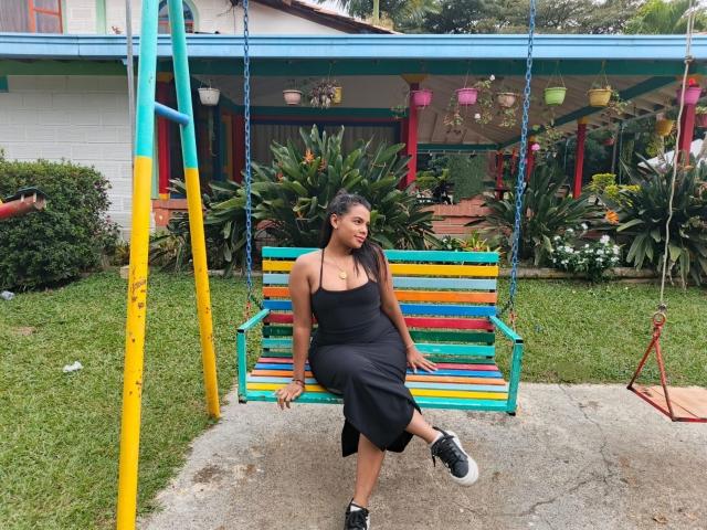 LissReyess sits on a colorful wooden swing, wearing a black dress, showing her ass with one leg raised, black sneakers, long brown hair, and a necklace.