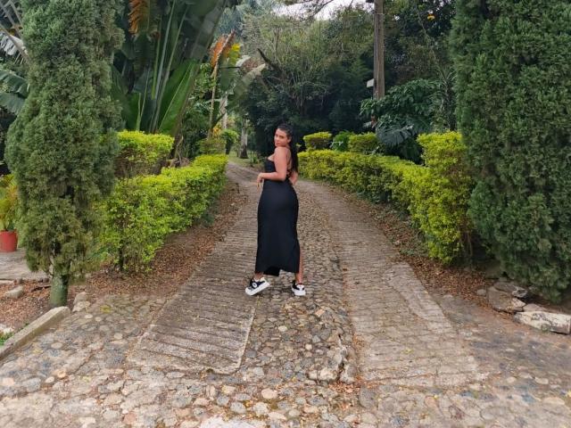LissReyess standing on a cobblestone path, black dress, sleeveless, black shoes with white soles, curvy ass, long dark hair, green bushes on both sides, forest background.