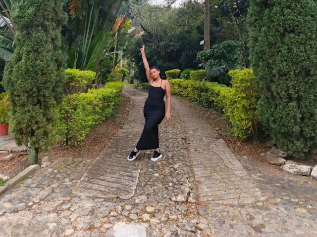 LissReyess, curvy with big ass, wearing a black strapless dress and sneakers, pointing up with one arm, standing on a cobblestone path, surrounded by greenery.