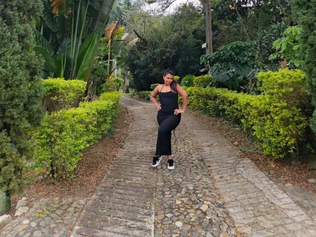 LissReyess stands on a cobblestone path wearing a black tank top and pants, with hands on her hips, and black sneakers, surrounded by green bushes and trees.