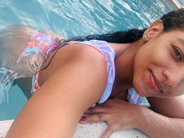 NatalyBlonder in a pool, wearing a colorful bikini, wet skin, and a smile, with her ass above the water, and her hand on the pool edge.