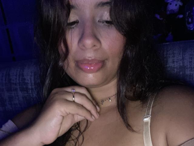 KeyklinOne with shiny pink lips, dark wavy hair, and a cute ring on her finger, wearing a beige top, has her eyes closed and a small smile. Blue light.