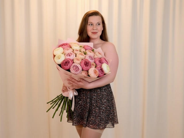 KarolynGlam stands in the center, holding a bouquet of pink and white roses with green stems. She has fair skin, long brown hair, and a subtle smile. She wears a floral skirt and is topless, with her arms gently holding the bouquet. The background is a plain, beige curtain. The image focuses on her and the bouquet, with soft, natural lighting highlighting her features. Her confident, relaxed pose contrasts with the delicate flowers. The composition is simple and centered.