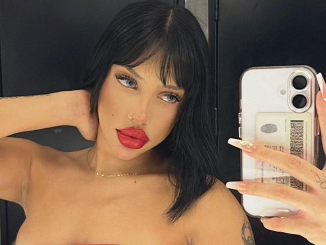 MelanieCollins with black hair, red lipstick, and long nails, taking a selfie with her phone, showing her bare boobs and tattooed arm.