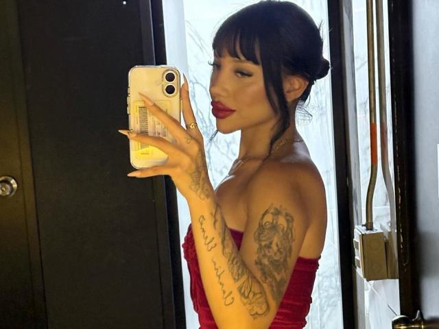 MelanieCollins with tattoos on her arm, wearing a red strapless dress, holding a phone, taking a mirror selfie, looking at the camera, red lipstick, dark hair in a bun.