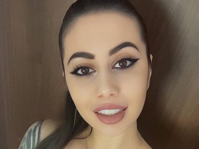 DiamondHottieX has a close-up selfie with a wooden background. She has dark brown hair pulled back, thick black eyebrows, and large brown eyes with black winged eyeliner. Her full pink lips are slightly parted, revealing white teeth. She wears small hoop earrings and a striped top is visible on the left. The image has a soft, natural light. The texture of her skin is smooth, and her makeup is bold and defined. Her hair is straight and sleek. The overall composition is centered and focused on her face.