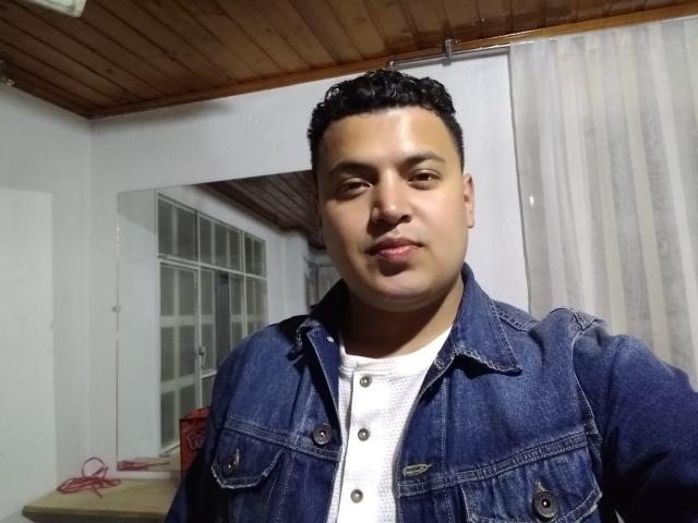 TheManagers has a medium build, short curly black hair, and a slight smile. He's wearing a blue denim jacket over a white shirt. His ass is not visible.