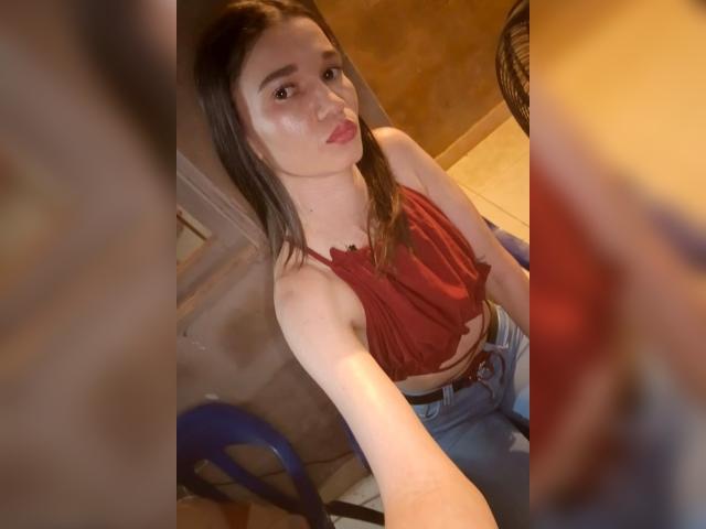 JennaDerose, con piel pálida, ojos marrones y cabello largo castaño, vestida con un top rojo de tirantes y jeans azules, tomando una selfie, mostrando sus pequeños senos y un maquillaje sutil..