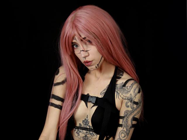 OliviaMonroee with pink hair, black-striped tattoos, and black bra, has a nose ring and dark makeup, looking seductive with a slight pout. Boobs visible, black background.