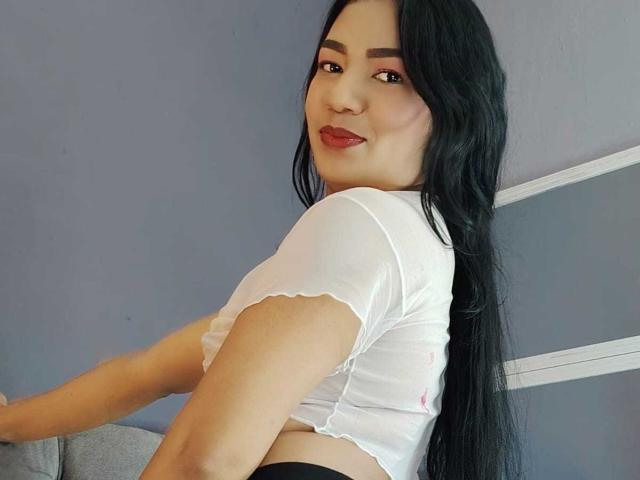 AnaPorto with long black hair, white sheer top, and red lipstick, looking back with a smile, showing her tits and black panties, in a selfie-style photo.