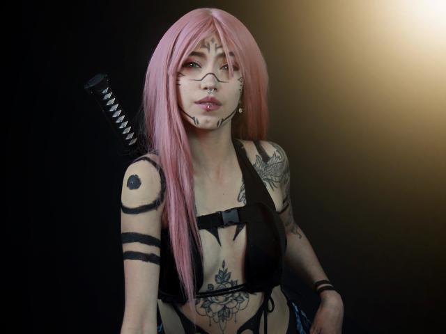 OliviaMonroee with pink hair, black bra, tattoos, black stripes on arms, sword on back, nose ring, and lip ring, looking at camera. Her tits are visible.