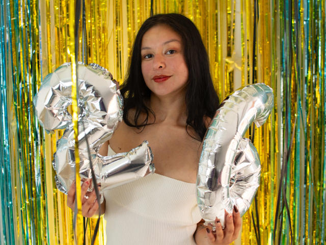 DyanaMoon with silver balloons in her hands, wearing a white strapless top, has long black hair and red lipstick, standing in front of shiny gold and green streamers.