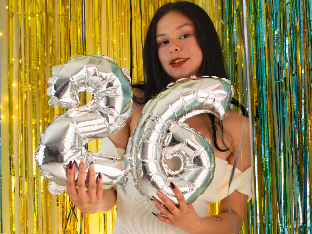 DyanaMoon sosteniendo globos plateados brillantes con la forma del número "26", con cabello negro, lápiz labial rojo y un vestido blanco, de pie frente a serpentinas metálicas amarillas y verdes. Sus manos están sobre los globos..