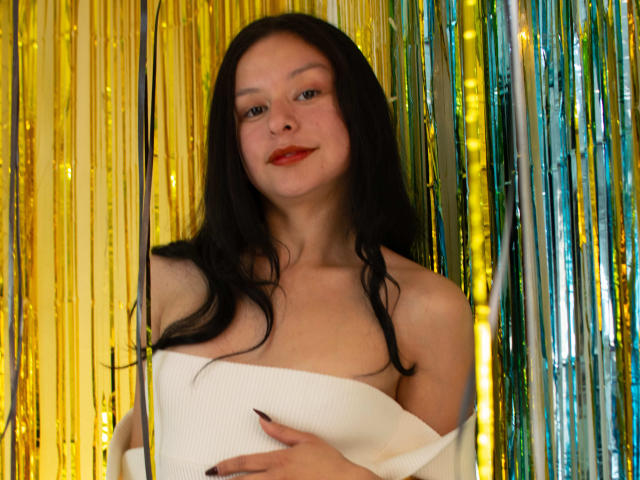 DyanaMoon, con cabello negro largo, lápiz labial rojo y una blusa blanca con un hombro descubierto, sosteniendo sus senos, rodeada de brillantes serpentinas doradas y verdes. Está mirando a la cámara..