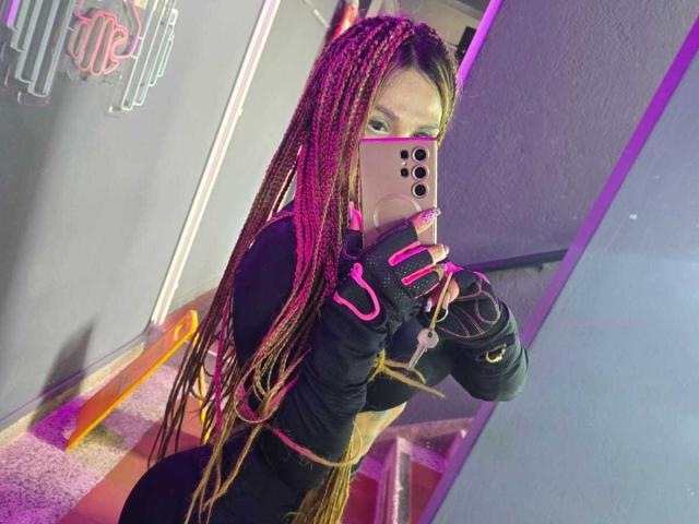 AzhunaStar with long pink and brown braids, black long-sleeve top, pink fingerless gloves, holding pink iPhone, taking mirror selfie in purple-lit room.