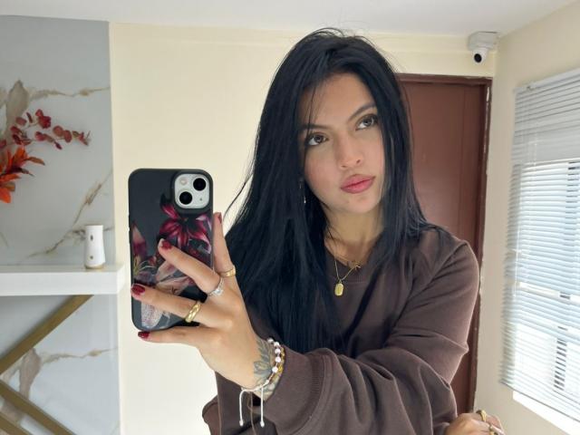 NyraVanel mit langen, schwarzen Haaren, braunen Augen und rosafarbenem Lippenstift, trägt einen braunen Pullover und macht ein Selfie mit einer Handyhülle mit Blumenmuster. Dabei zeigt sie ihre roten Nägel und ihren goldenen Schmuck..