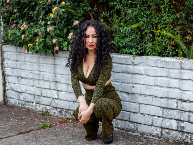 SamanthaAntolin crouches on a concrete surface in front of a white brick wall. She has curly black hair, red lipstick, and wears an olive green knitted crop top and pants. Her top reveals a deep cleavage. She has a medium build with a curvy figure. The background is lush with green foliage and pink flowers. She is looking slightly to the left with a subtle, confident smile. Her black heels are visible. The image has a natural, candid feel.