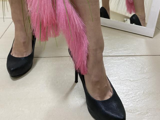 Amberia's legs in black high heels with pink feather accents, standing on a tiled floor. Her reflection is visible in a mirror behind her.
