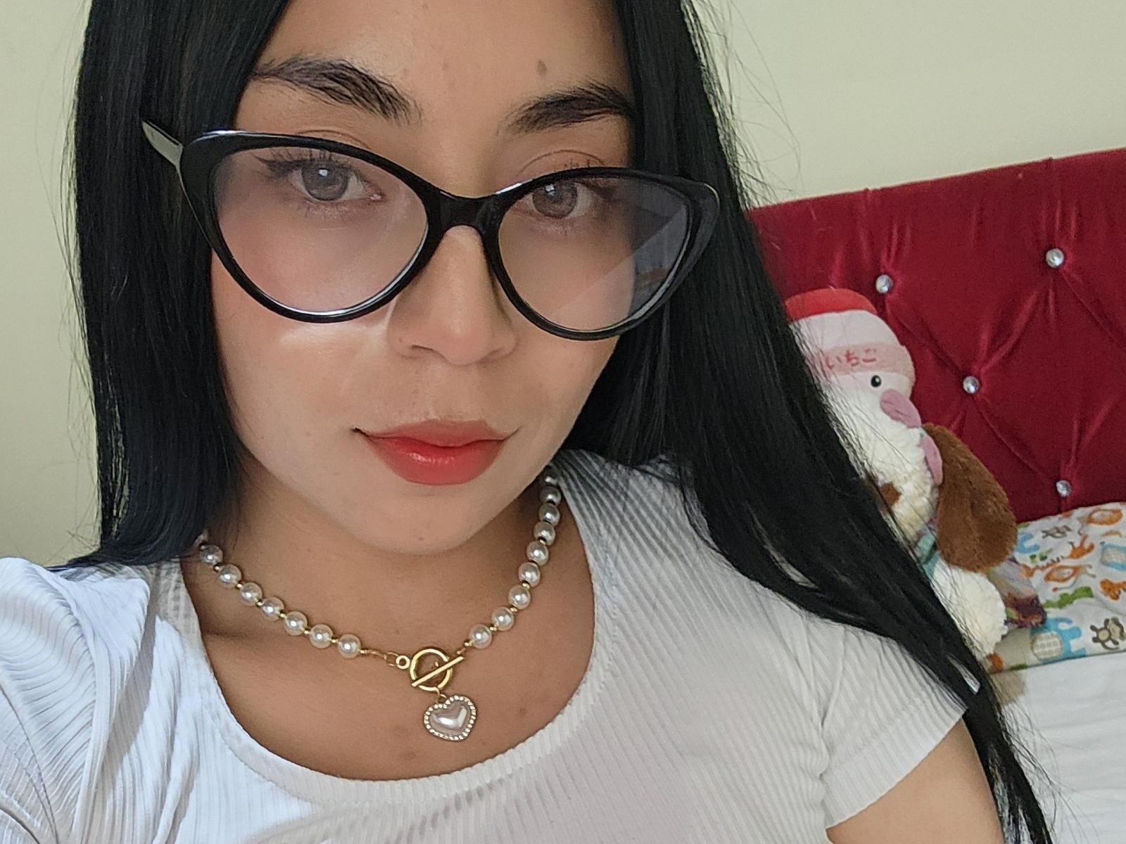 Luisianna with black glasses, red lipstick, white ribbed shirt, pearl necklace with heart pendant, long black hair, looking at camera, red tufted headboard with stuffed Santa.