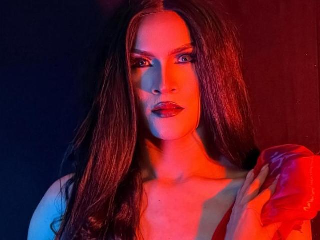 AgathaRodriguez has long, wet-looking brown hair, illuminated by red and blue lighting. She holds a red, puffy fabric in her right hand. Her makeup is bold, with dark red lipstick and dramatic eye shadow. Her skin is light, and her expression is serious. The red and blue lights create stark contrasts, highlighting her facial features and hair texture. The image is highly saturated, emphasizing the contrasting colors.