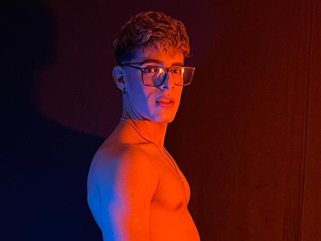 Shirtless AngelEvil with glasses, short curly blonde hair, and a necklace, illuminated by blue and red lights, looking over shoulder with a slight smile.